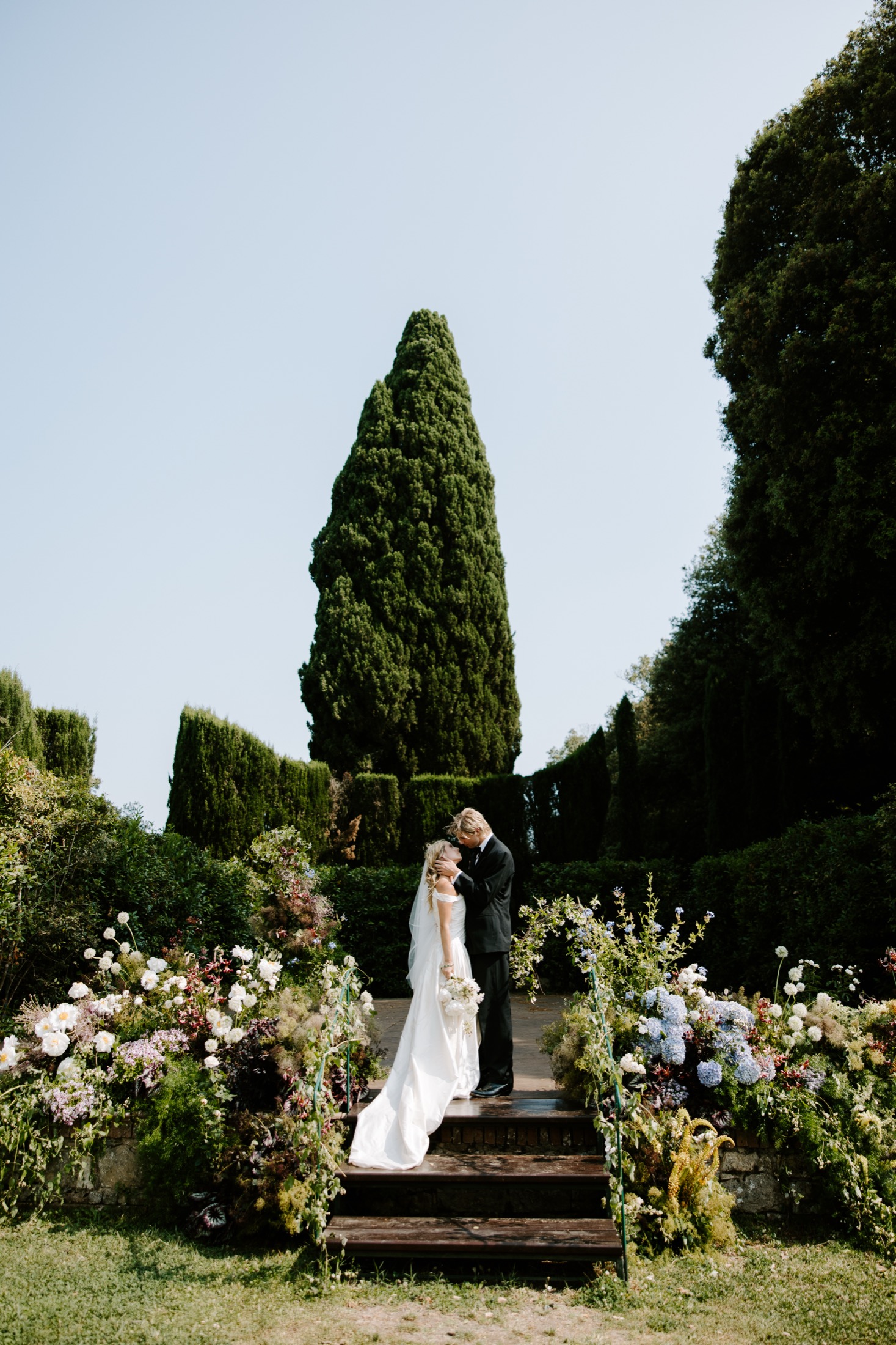 Bride and groom kissing among flowers and cypress trees in an Italian garden