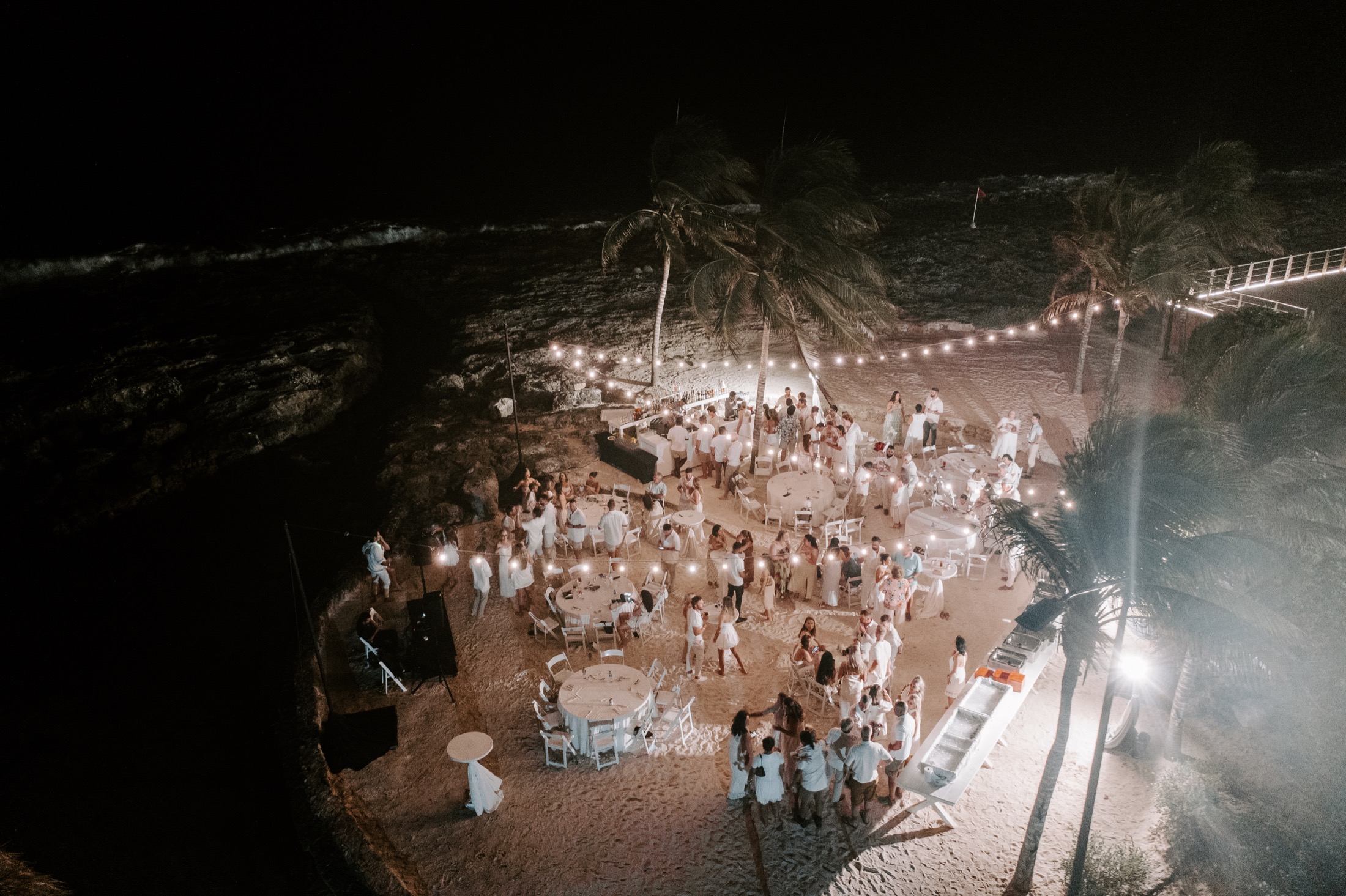 Aerial view of a wedding reception beneath string lights beside the beach
